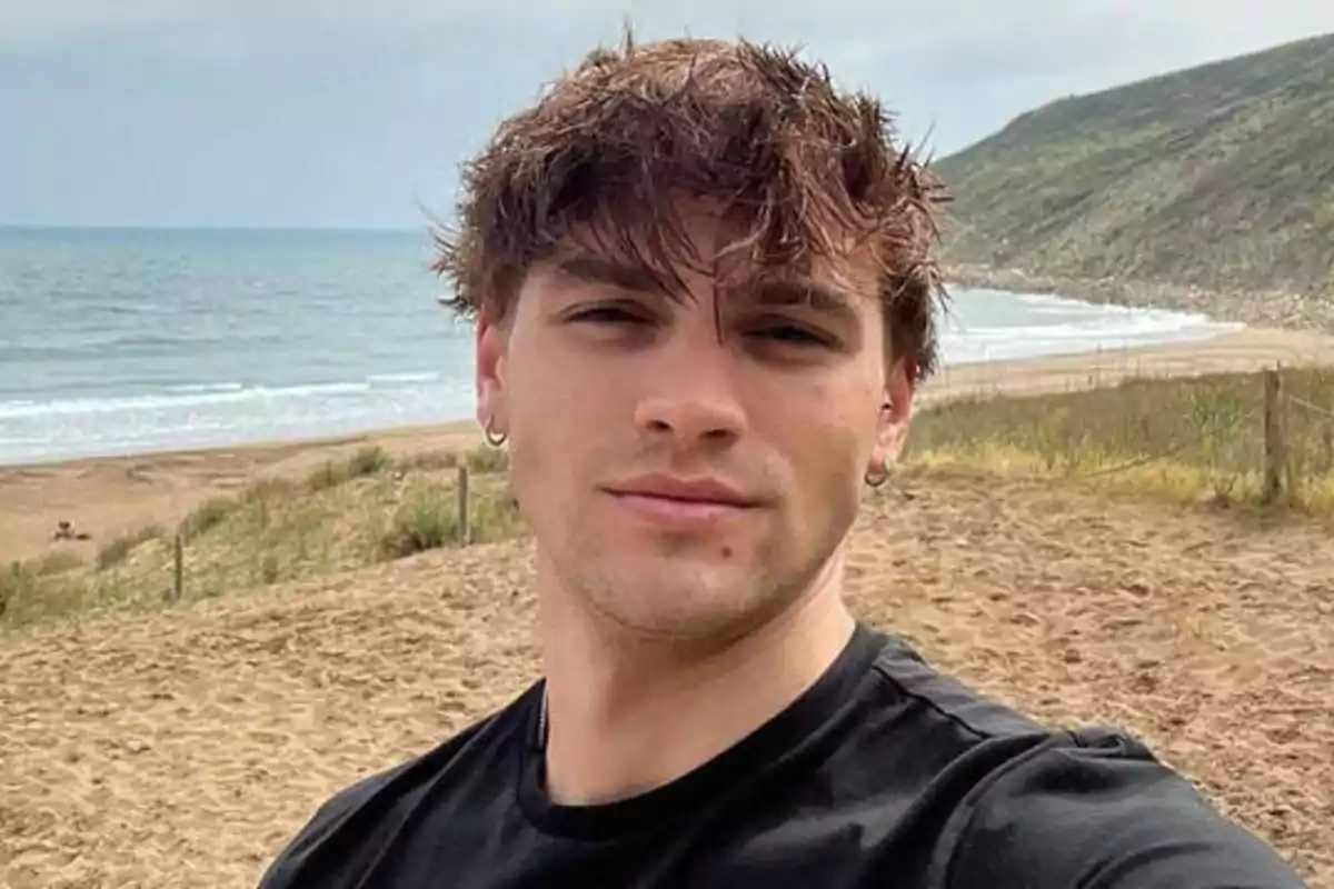 Un hombre joven con cabello despeinado y camiseta negra posando para una selfie en una playa con el mar y colinas al fondo.