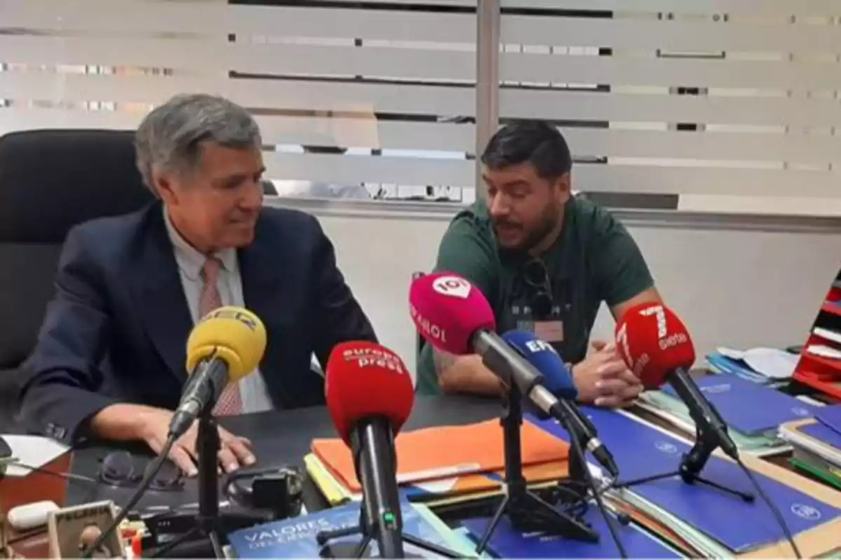 Dos hombres sentados en una mesa con varios micrófonos de diferentes medios de comunicación frente a ellos.