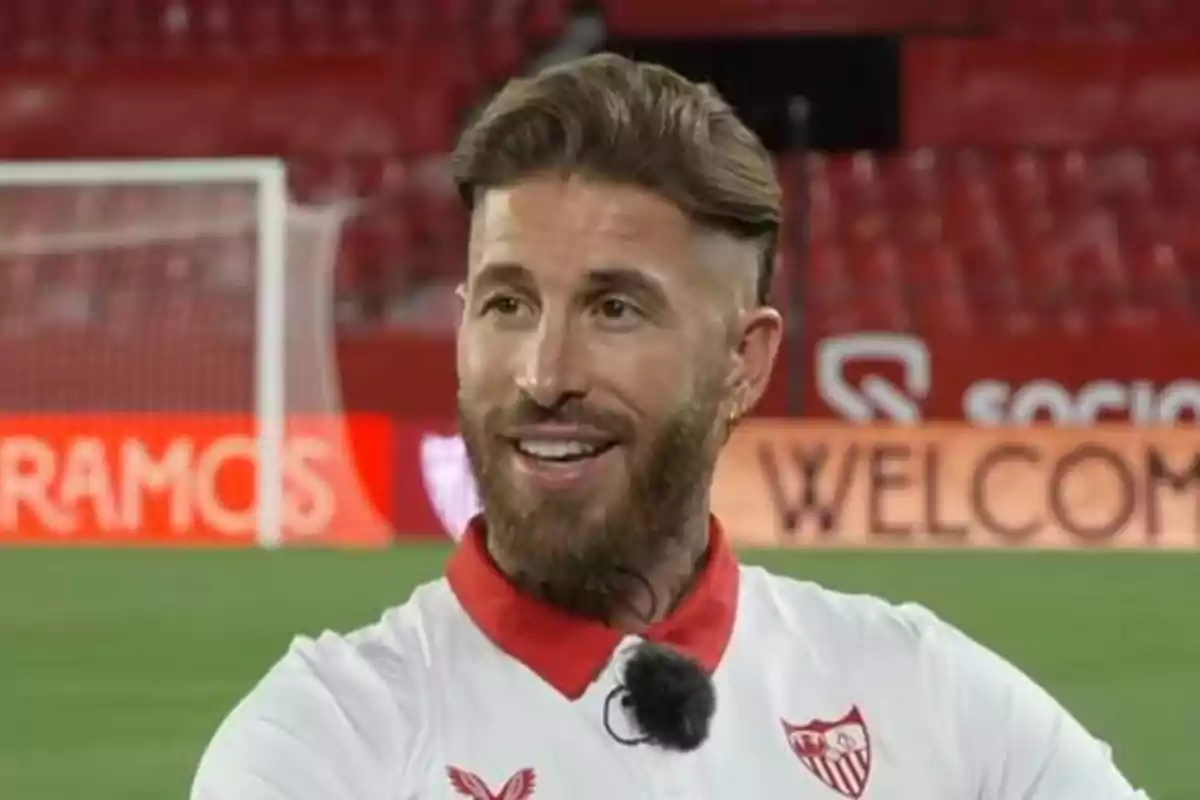 Un jugador de fútbol con uniforme blanco y rojo del Sevilla FC sonríe en un estadio.