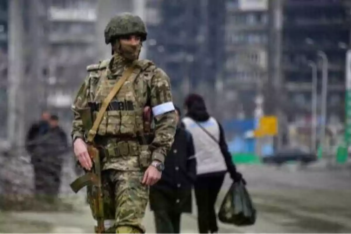 Un soldado con uniforme de camuflaje y casco camina por una calle en una zona urbana, mientras dos personas pasan por detrás.