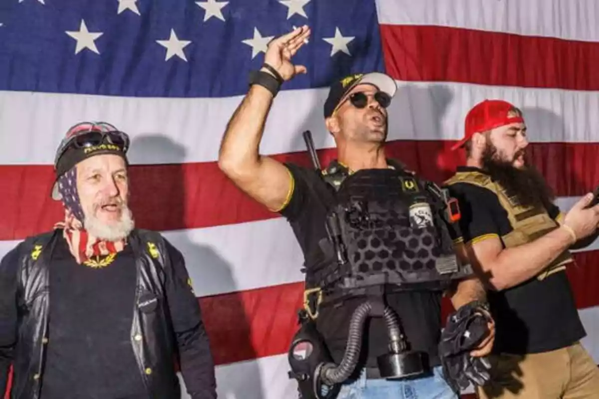 Tres hombres con vestimenta militar y accesorios están de pie frente a una bandera de Estados Unidos.