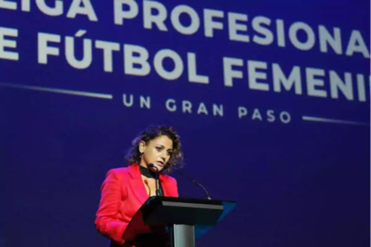 Una mujer con chaqueta roja hablando en un podio frente a un fondo que dice "LIGA PROFESIONAL DE FÚTBOL FEMENINO UN GRAN PASO".