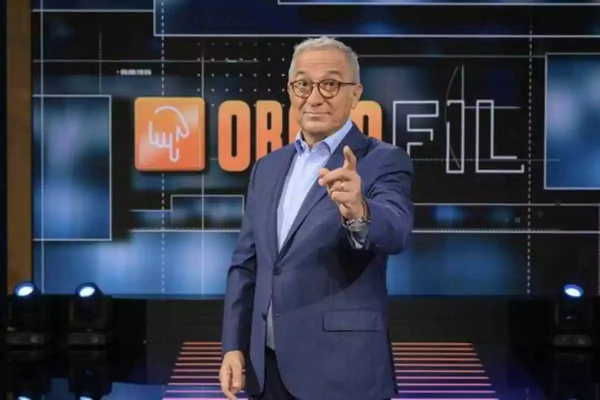 Un hombre con gafas y traje azul señala hacia la cámara mientras está de pie frente a un fondo con el logotipo de un programa de televisión.