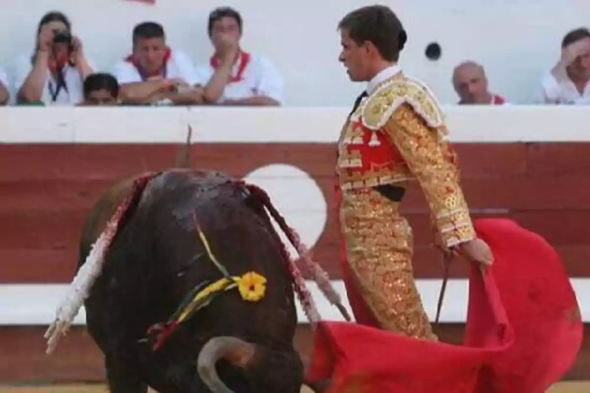 Un torero vestido con traje de luces dorado sostiene una muleta roja frente a un toro en una plaza de toros, mientras el público observa desde las gradas.