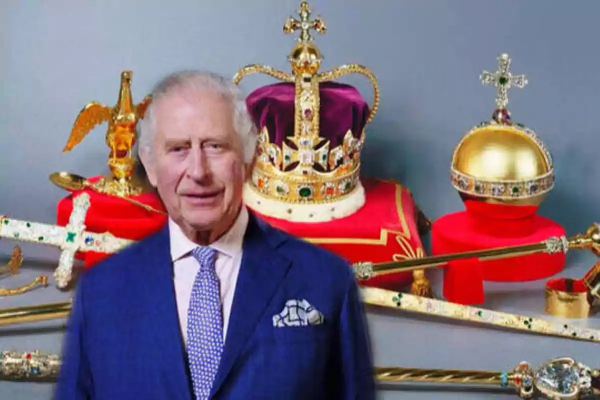 Un hombre mayor con traje azul y corbata clara está de pie frente a una colección de joyas de la corona, incluyendo una corona, un orbe y varios cetros.