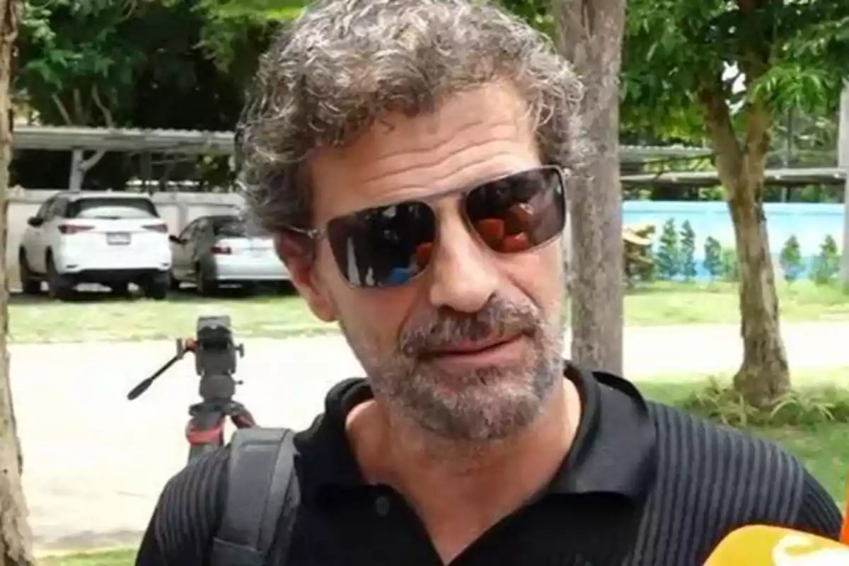 Un hombre con gafas de sol y barba está al aire libre, con árboles y coches estacionados en el fondo.