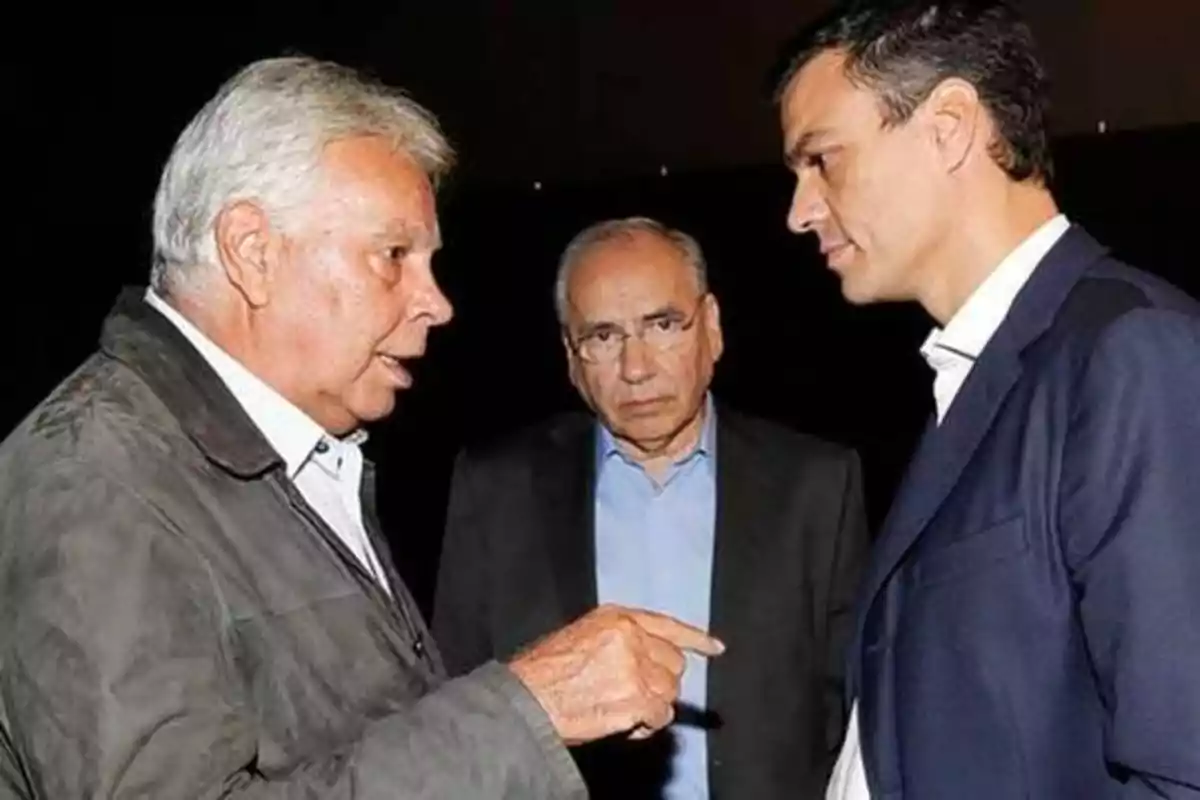 Tres hombres conversando en un entorno oscuro, uno de ellos gesticulando con la mano.