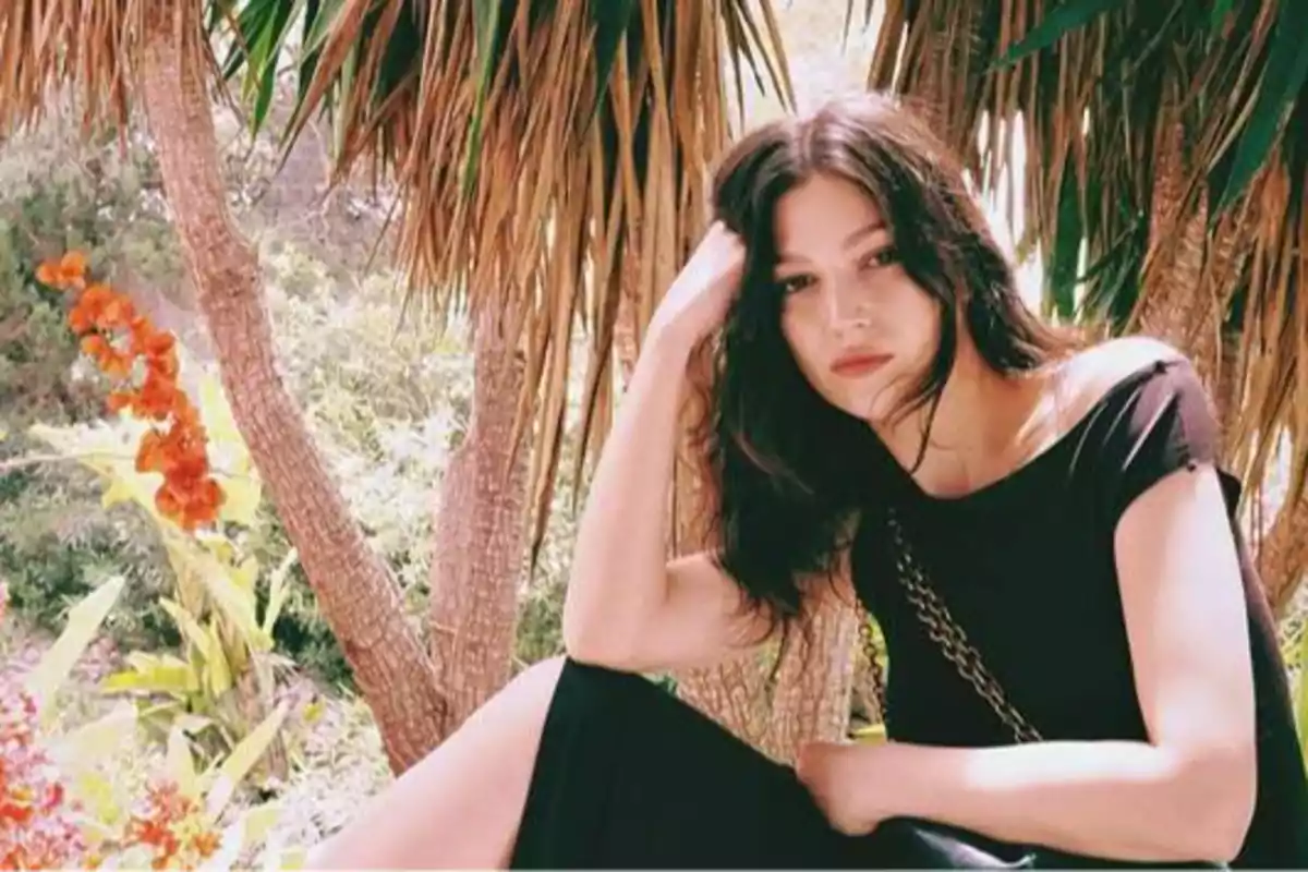 Mujer con vestido negro sentada bajo un árbol con hojas largas y verdes, rodeada de vegetación y flores rojas.