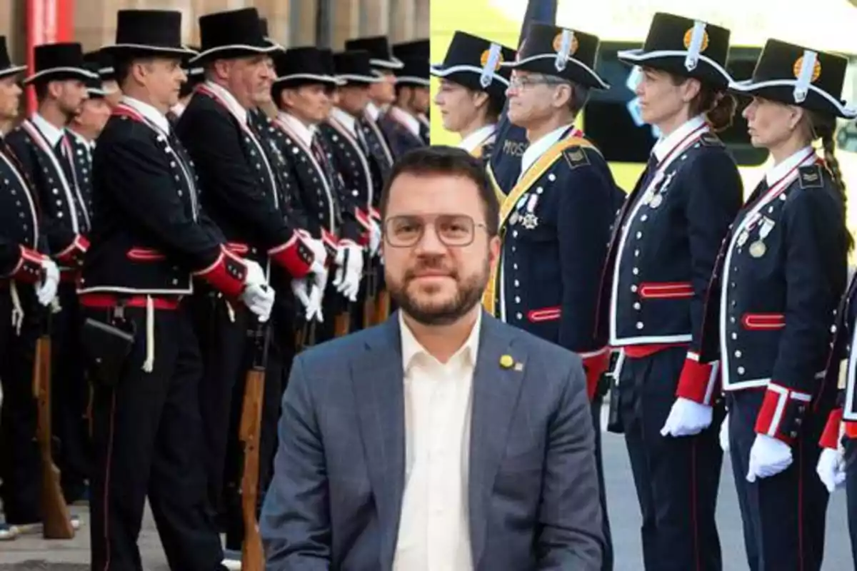 Un hombre con gafas y barba está sentado frente a dos filas de personas uniformadas con trajes formales y sombreros, una fila de hombres a la izquierda y una fila de mujeres a la derecha.