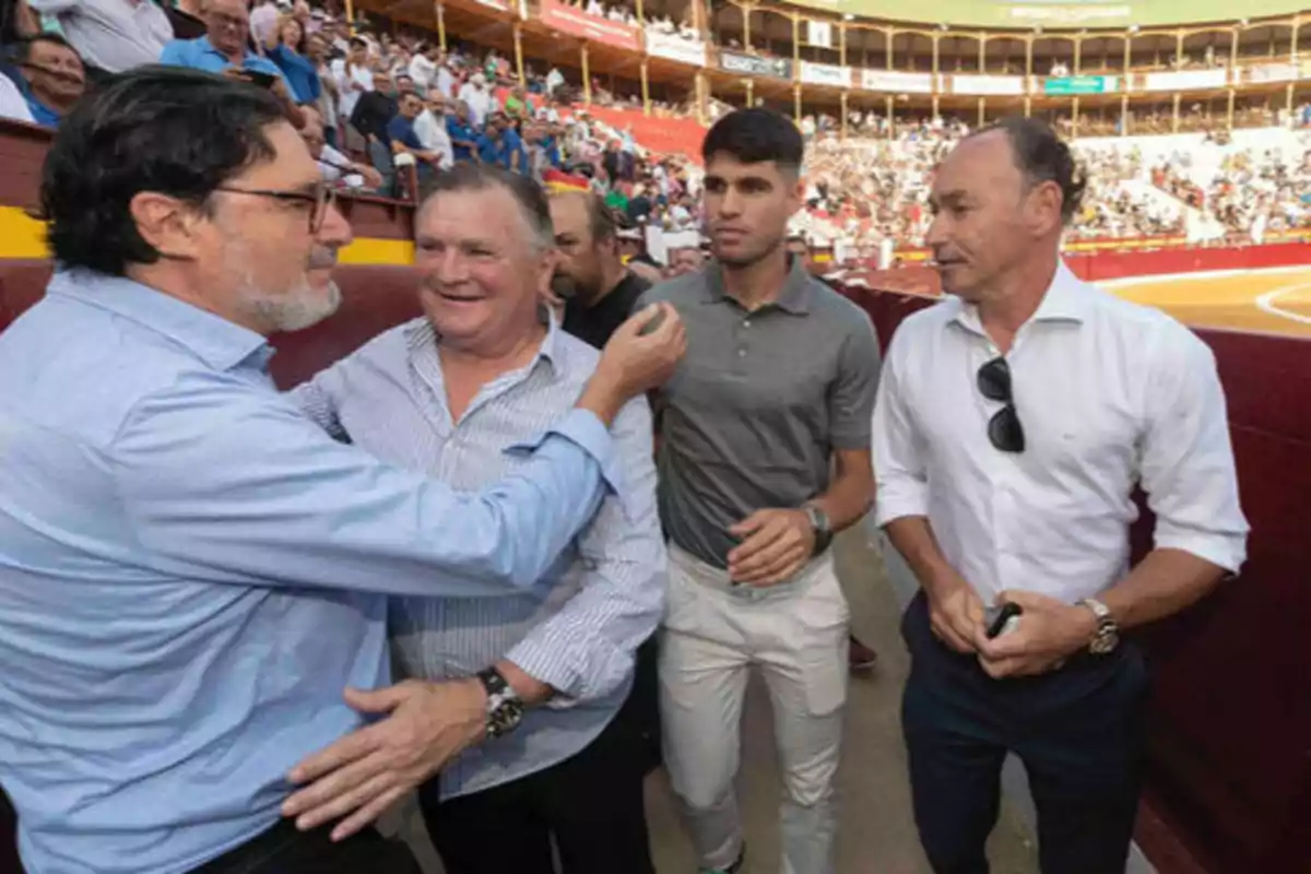 Un grupo de hombres se encuentra en una plaza de toros, conversando y sonriendo mientras la multitud observa desde las gradas.