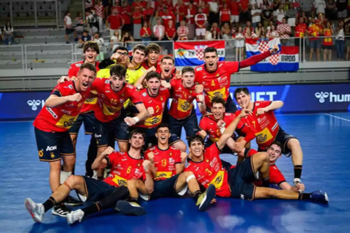 Un grupo de jugadores de balonmano con uniformes rojos y amarillos celebra en una cancha cubierta, con una multitud de seguidores en las gradas al fondo.