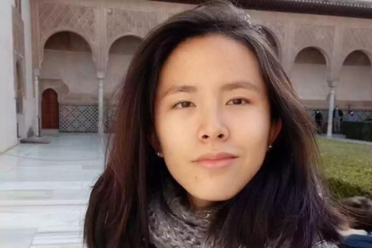 Una mujer con cabello largo y oscuro, usando una bufanda gris, está de pie frente a un edificio con arcos y detalles arquitectónicos intrincados.