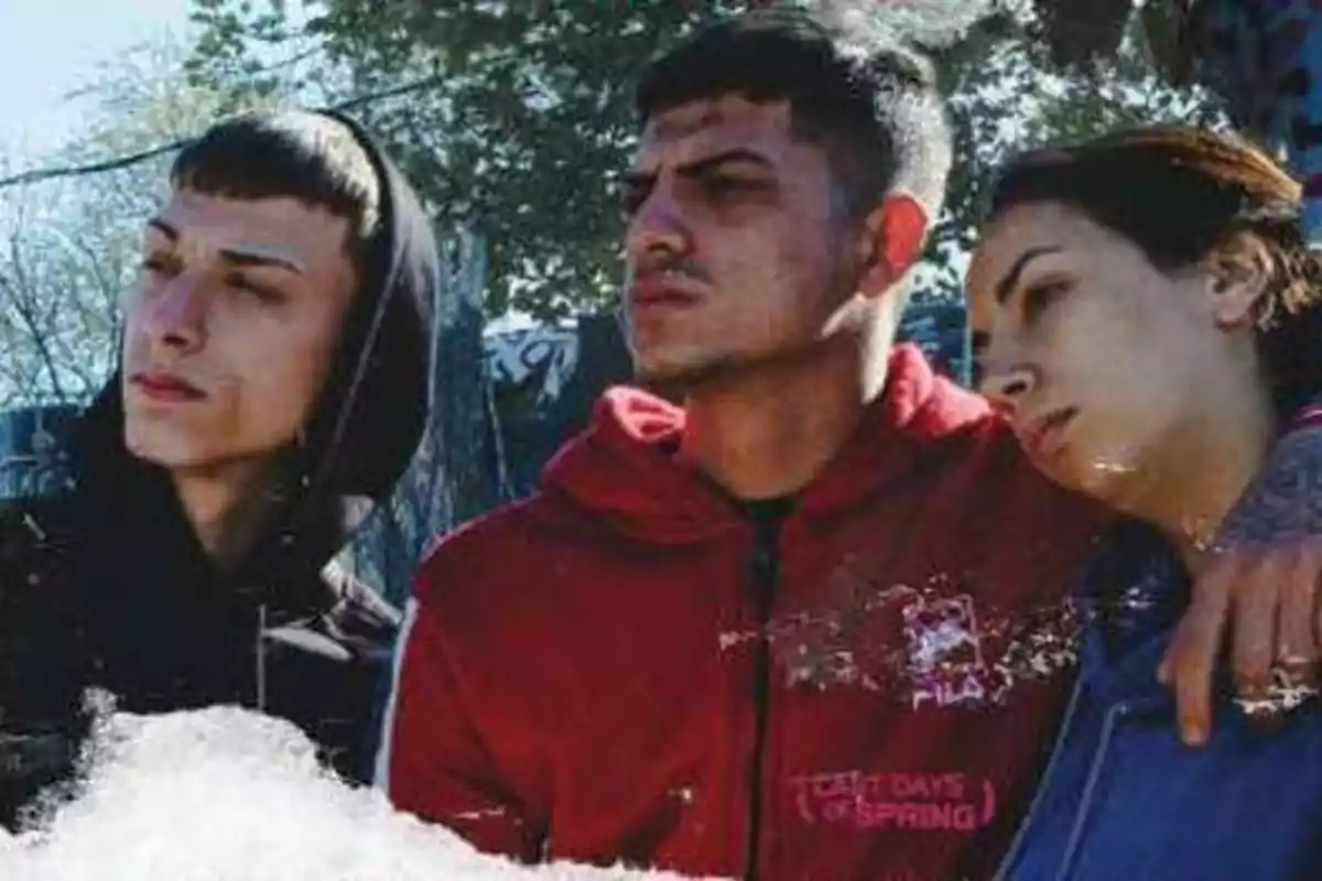 Tres jóvenes con sudaderas, uno con capucha negra, otro con sudadera roja y una chica con sudadera azul, posan juntos al aire libre.