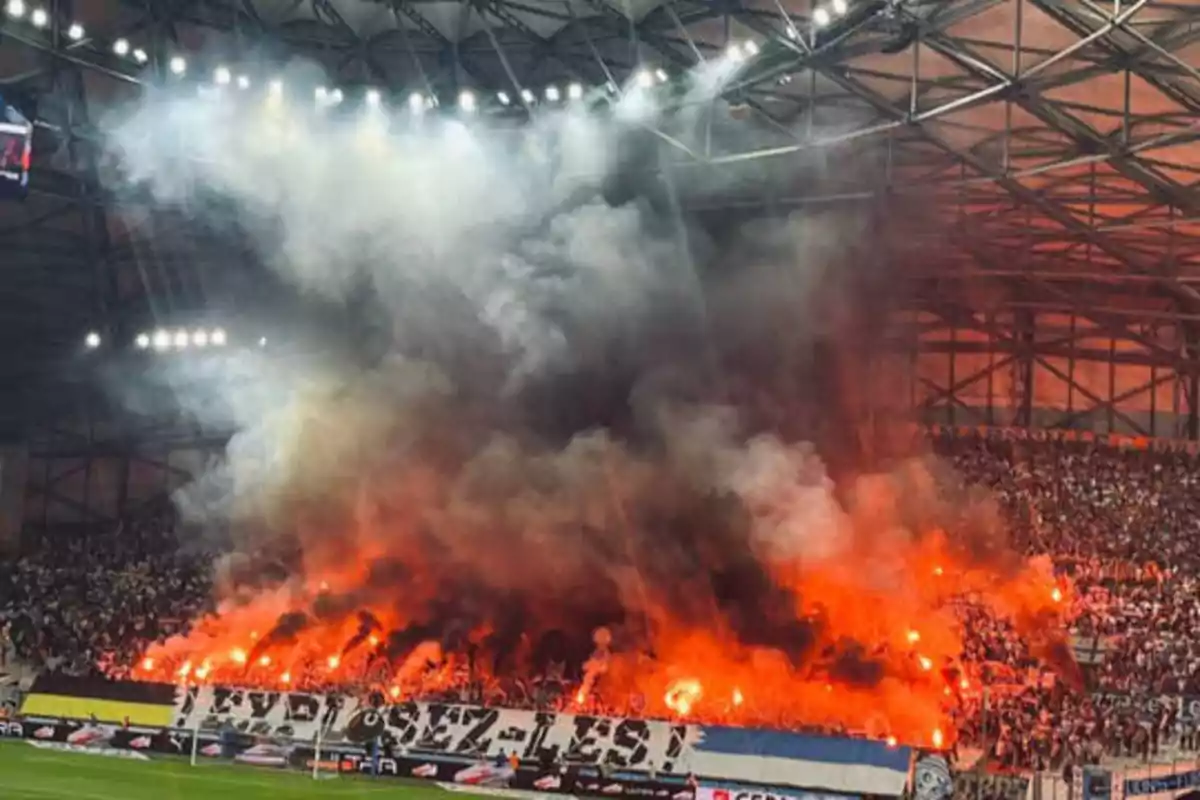 Aficionados en un estadio de fútbol encendiendo bengalas y generando una gran cantidad de humo.