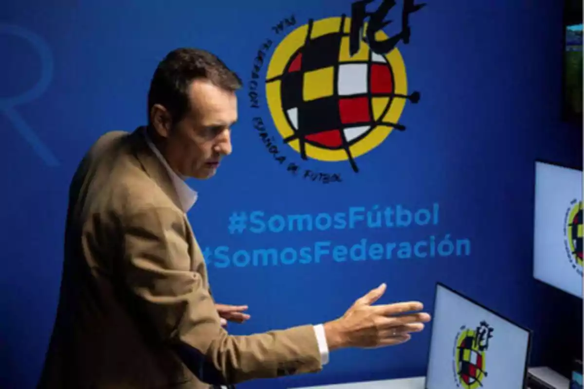 Un hombre de traje claro gesticula frente a una pantalla con el logo de la Real Federación Española de Fútbol y los hashtags #SomosFútbol y #SomosFederación en el fondo azul.