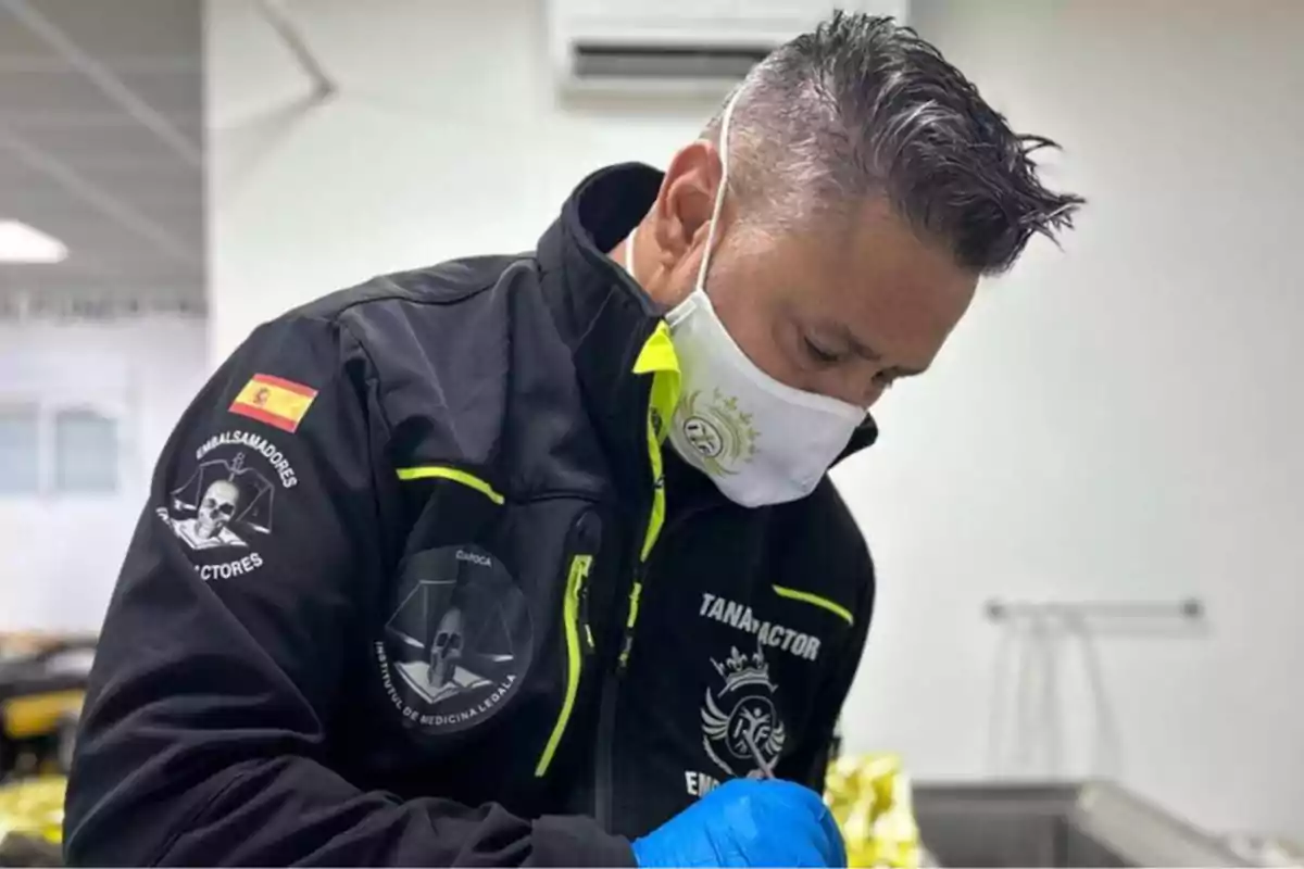 Un hombre con mascarilla y guantes azules trabaja en un entorno profesional con un uniforme que tiene insignias y una bandera de España.