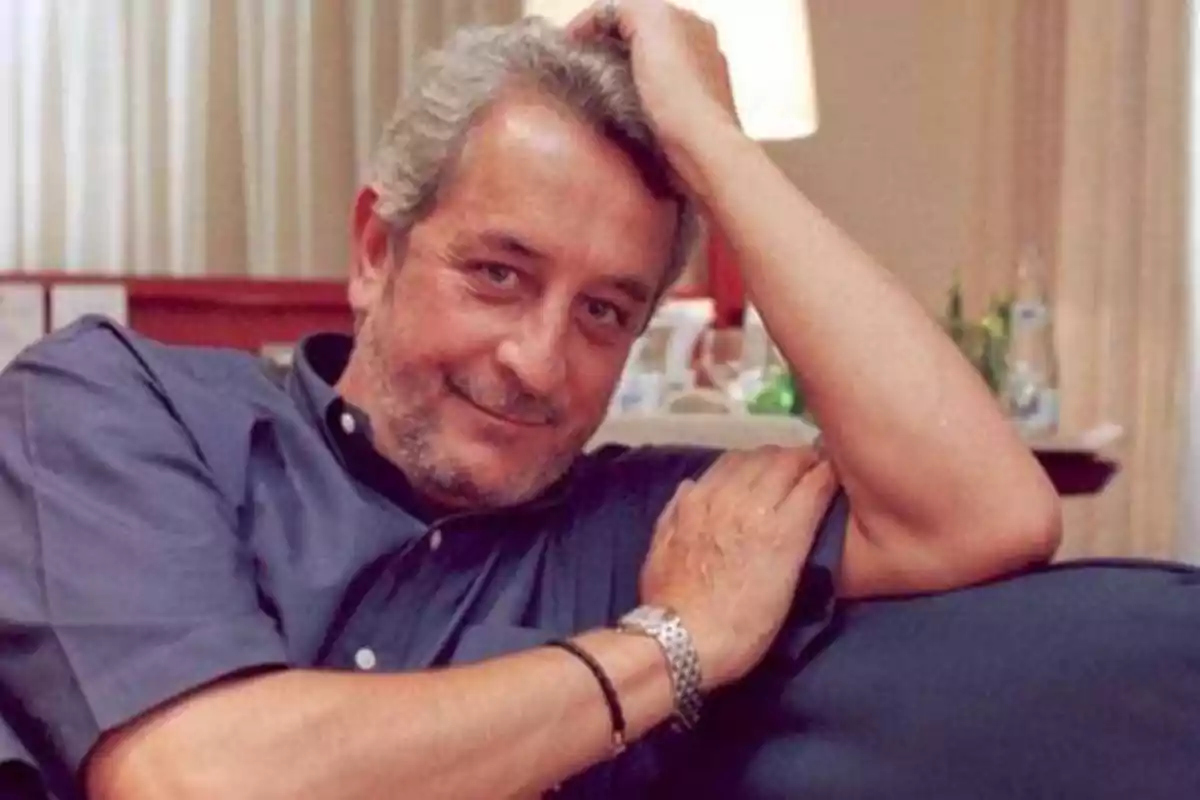 Hombre de cabello canoso y barba, con camisa azul, apoyado en un sofá y sonriendo.