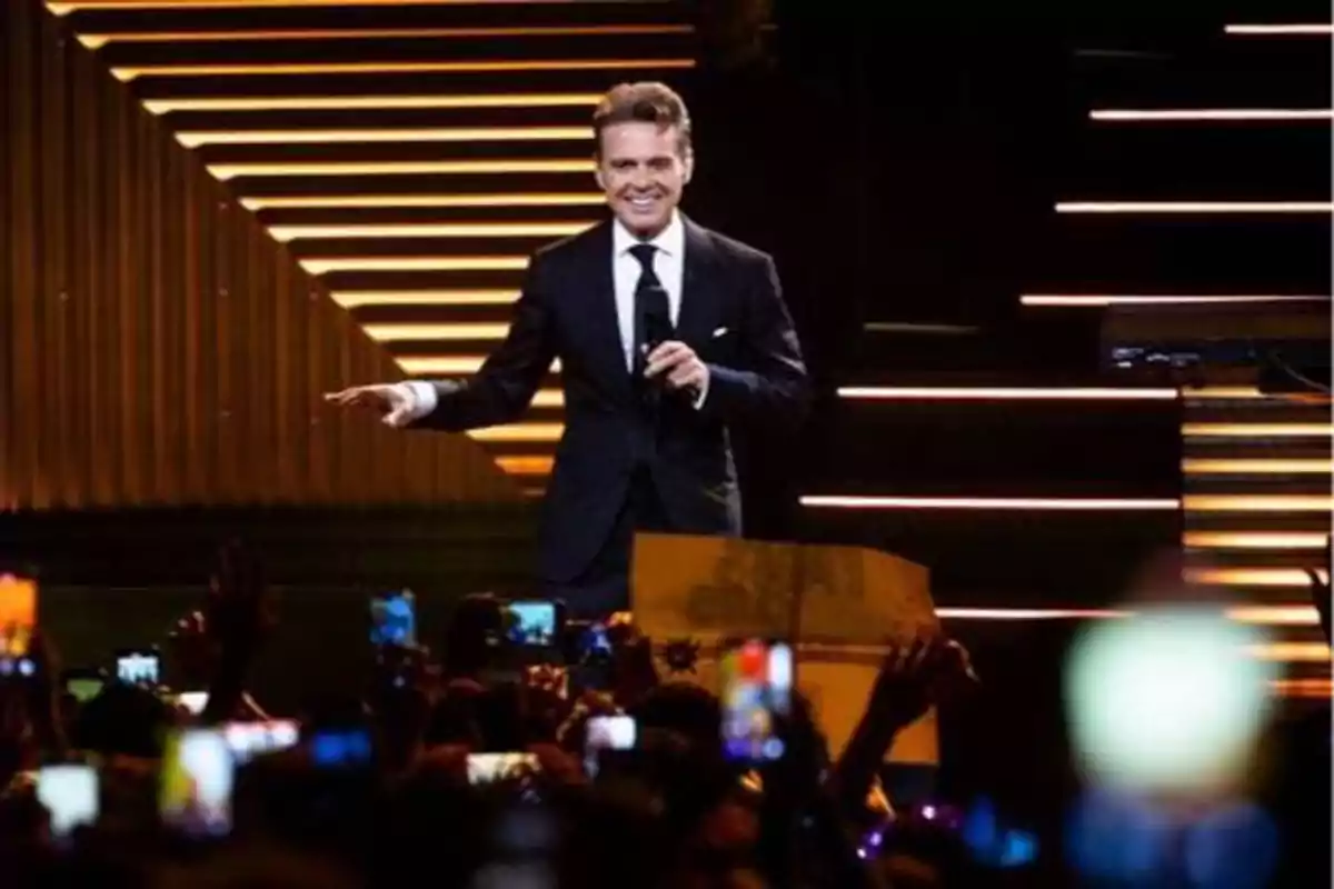 Un hombre en traje negro con micrófono en mano, sonriendo y saludando a una multitud que lo fotografía con sus teléfonos, en un escenario iluminado con luces y escaleras.