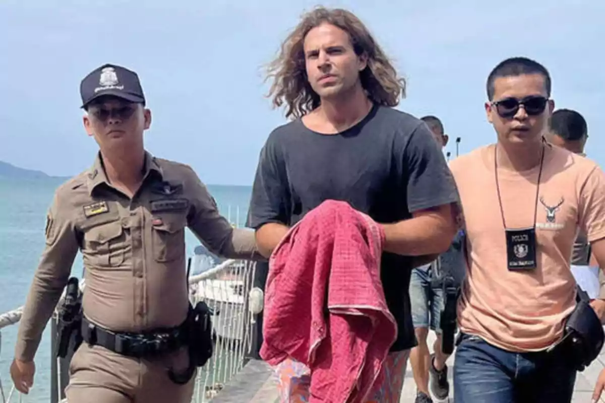 Un hombre con una camiseta negra y pantalones cortos coloridos está siendo escoltado por dos oficiales de policía, uno a cada lado, mientras camina por un muelle con el mar de fondo.