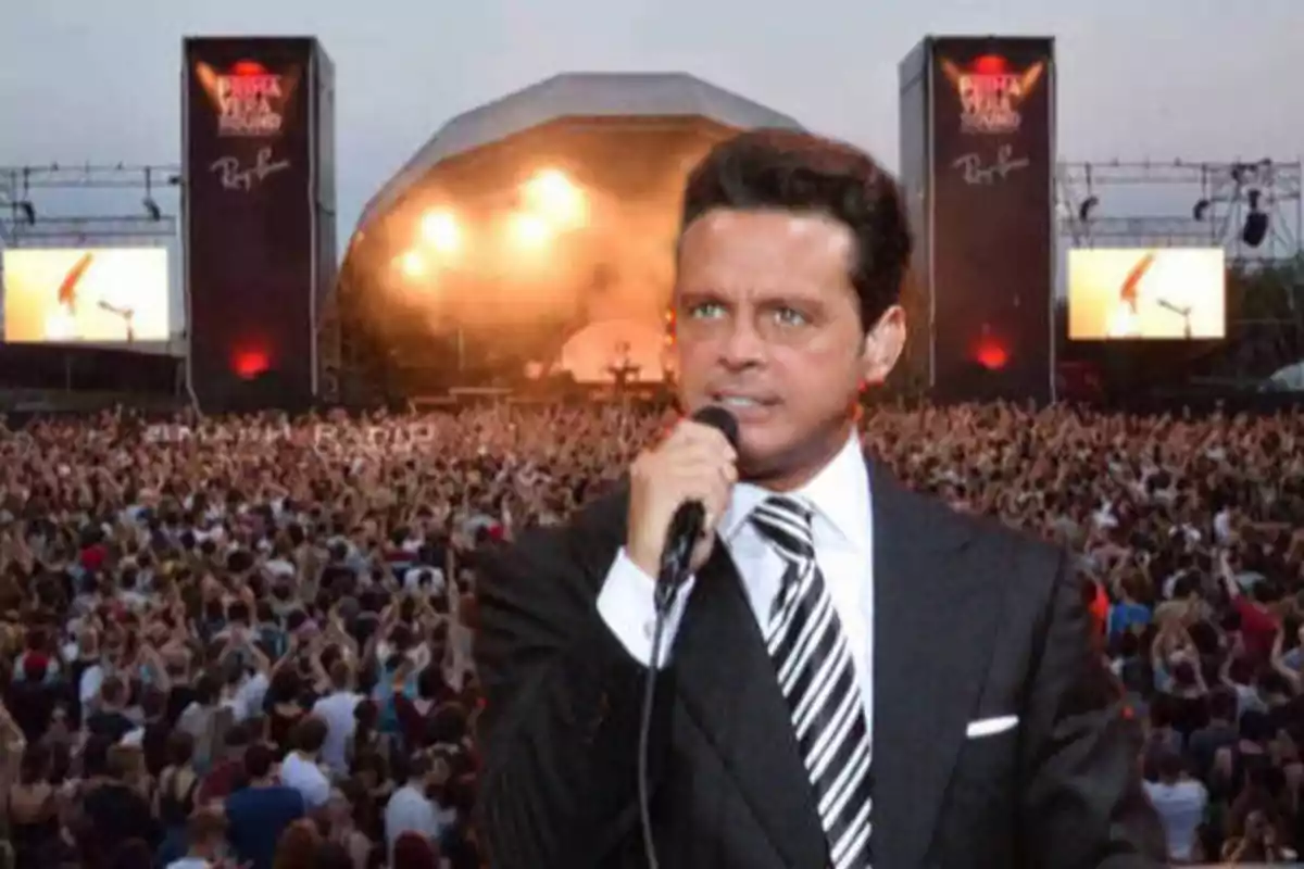 Un hombre con traje y corbata sostiene un micrófono frente a una multitud en un concierto al aire libre con un escenario iluminado al fondo.