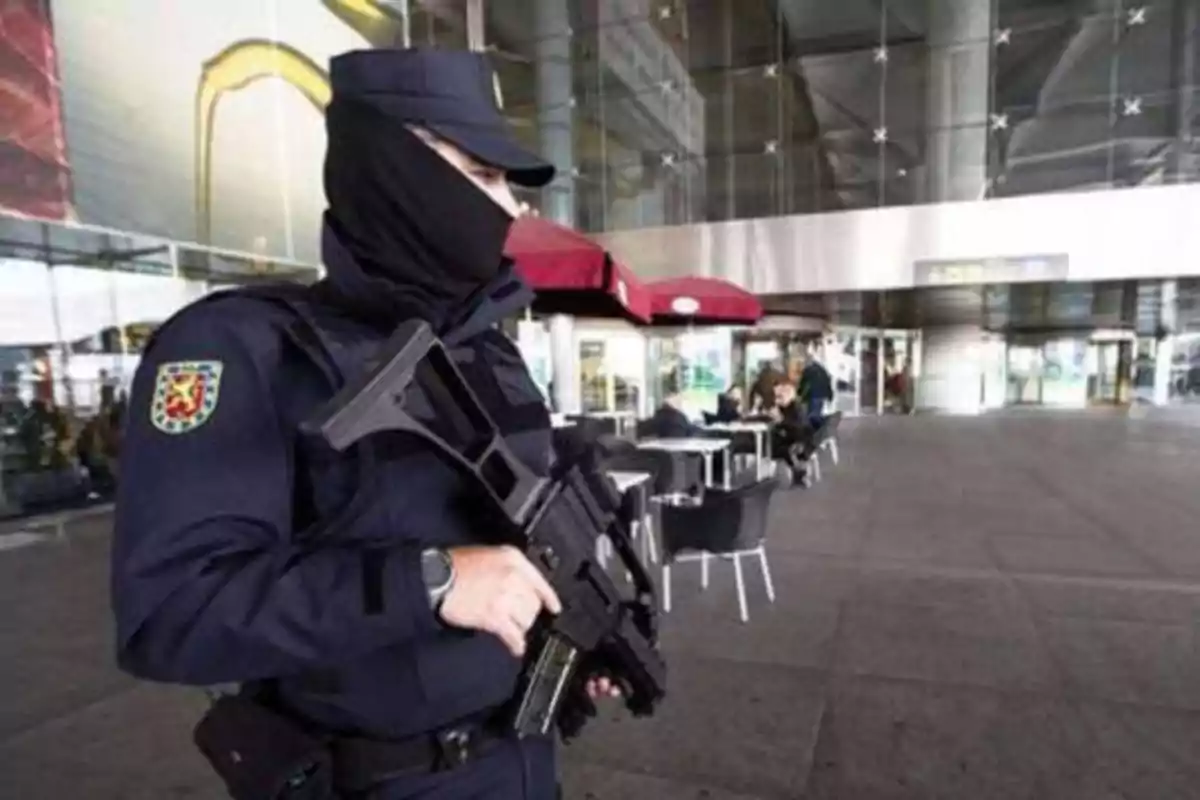Policía con uniforme y pasamontañas sosteniendo un arma frente a un edificio con mesas y sillas al aire libre. Policía con uniforme y pasamontañas sosteniendo un arma frente a un edificio con mesas y sillas al aire libre.