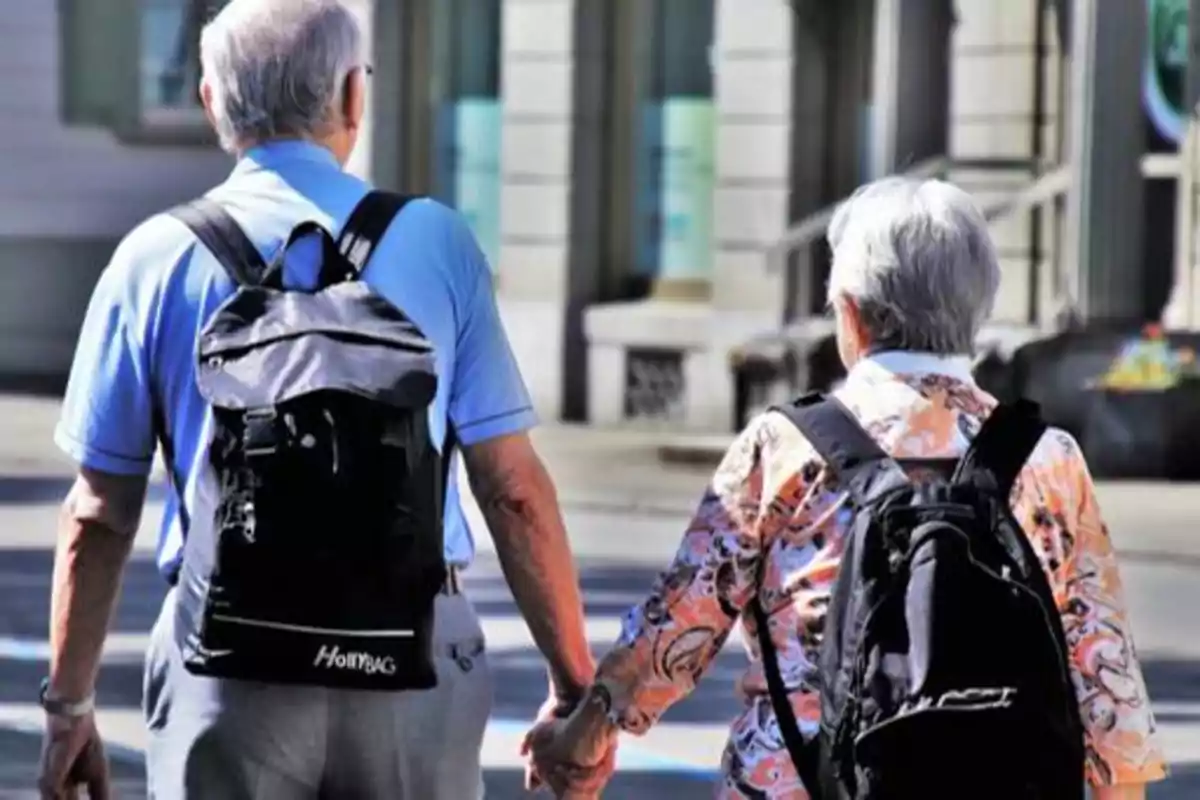 Dos personas mayores caminando de la mano con mochilas en la espalda.