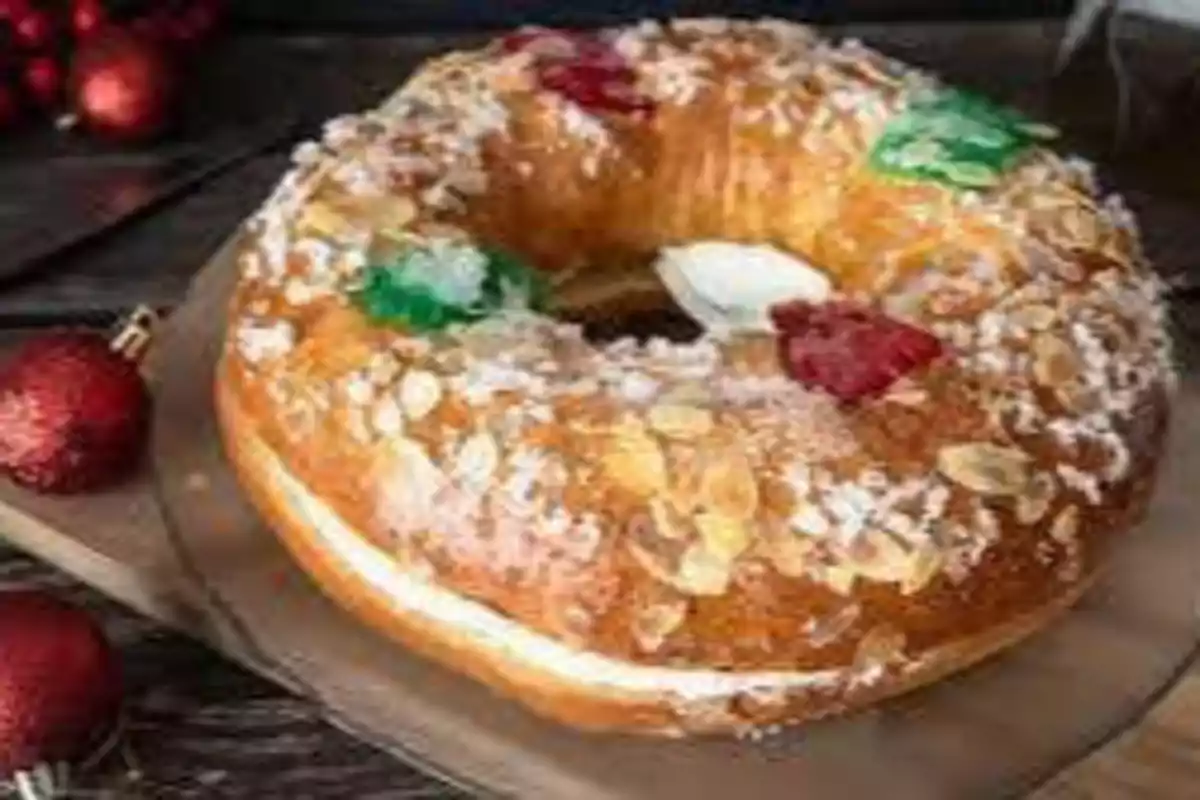 "Rosca de Reyes decorada con frutas cristalizadas y azúcar, colocada sobre una tabla de madera con esferas navideñas rojas al fondo."