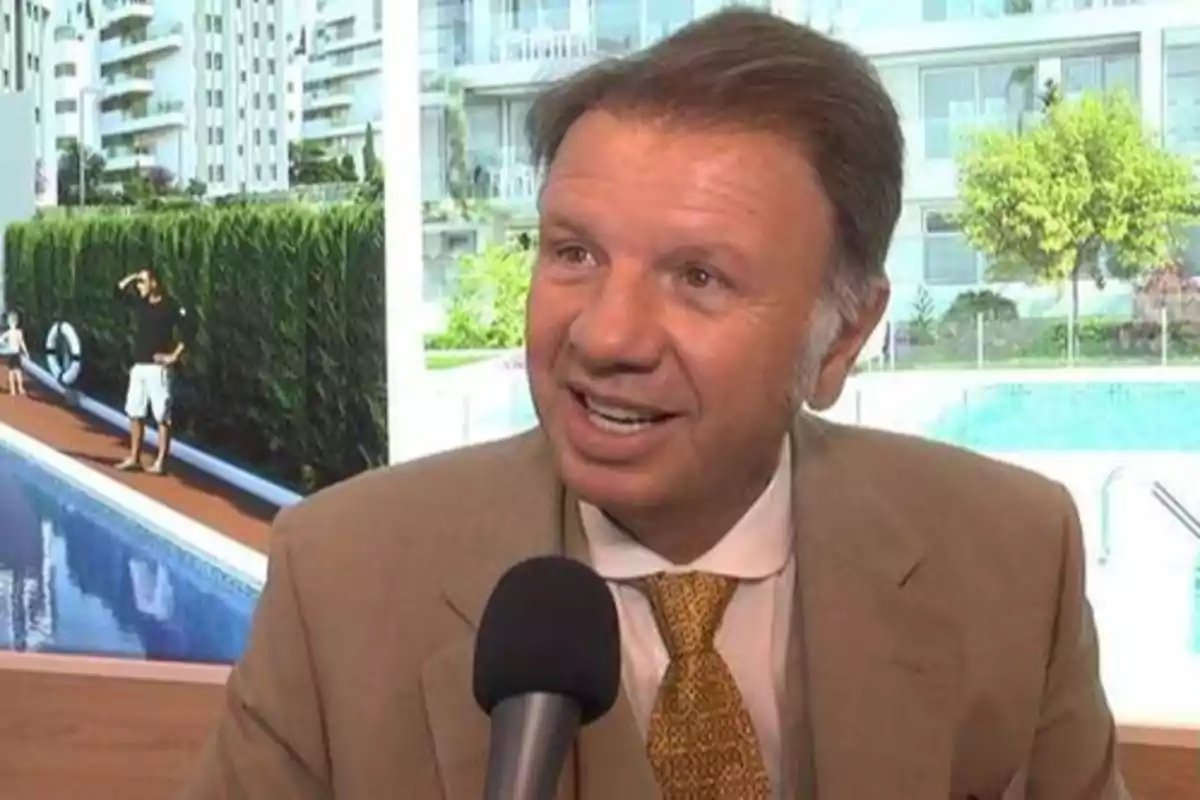 Hombre con traje y corbata dorada hablando frente a un micrófono con un fondo de edificios y una piscina.