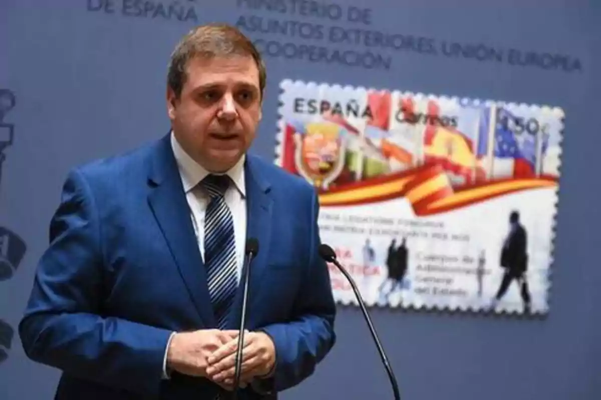 Un hombre con traje azul y corbata a rayas habla en un podio con dos micrófonos. Detrás de él, hay una imagen ampliada de un sello postal de España. En el fondo, se puede leer parcialmente el texto "MINISTERIO DE ASUNTOS EXTERIORES, UNIÓN EUROPEA Y COOPERACIÓN".
