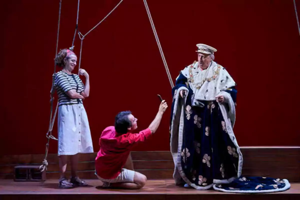 Tres personas en un escenario, una mujer de pie a la izquierda, un hombre arrodillado en el centro y otro hombre vestido con un traje real a la derecha.