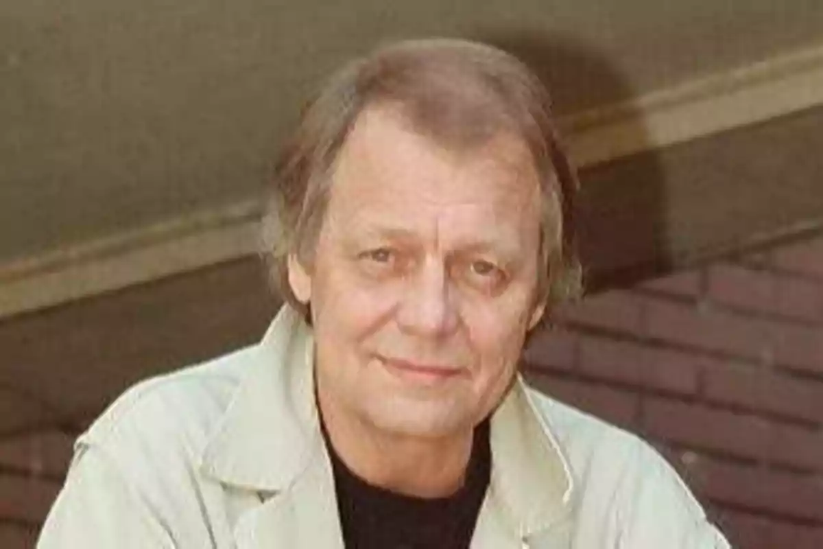 Un hombre mayor con cabello canoso y una expresión amable, vestido con una chaqueta clara, posando frente a un fondo de ladrillos.