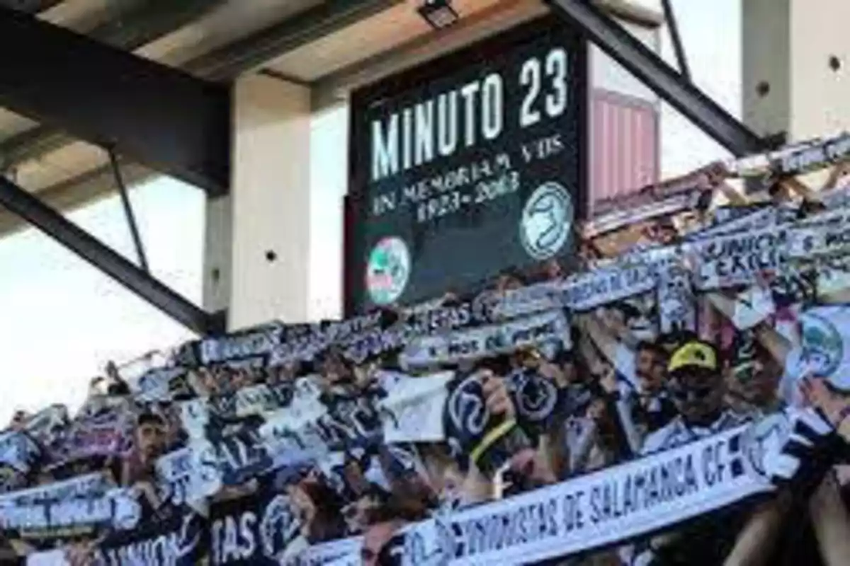 Aficionados en un estadio de fútbol levantando bufandas mientras un marcador muestra "Minuto 23" en memoria de una persona.