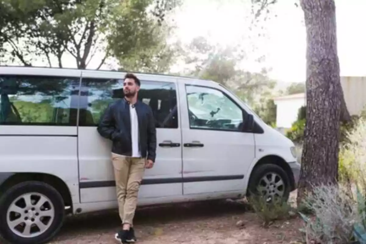 Hombre de pie junto a una furgoneta blanca en un entorno natural con árboles.