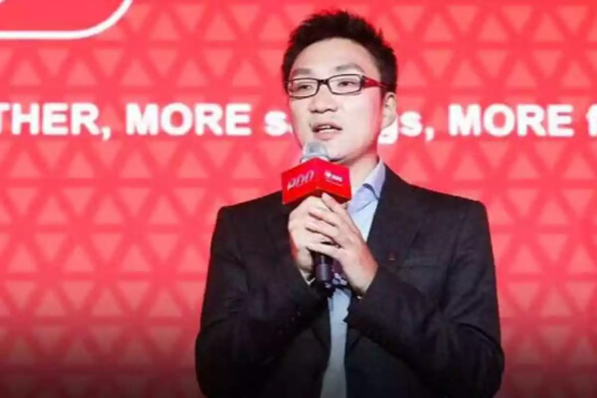 Hombre con gafas hablando en un micrófono frente a un fondo rojo con texto.