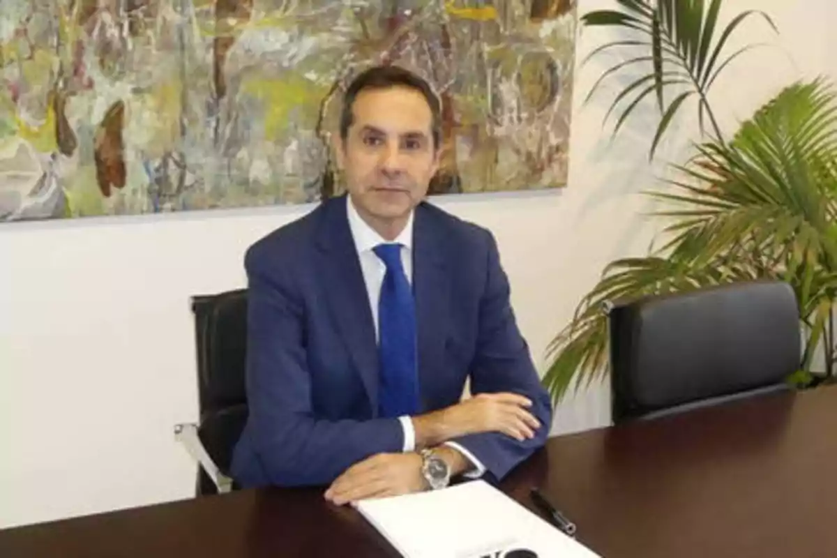 Un hombre con traje azul y corbata azul está sentado en una mesa de oficina con un bloc de notas y un bolígrafo, con una planta y una pintura abstracta en el fondo.