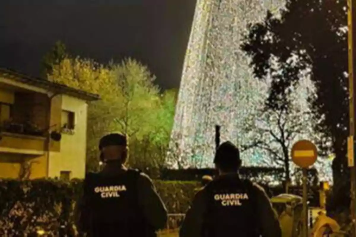 Dos agentes de la Guardia Civil de espaldas observan un árbol de Navidad iluminado en la noche.