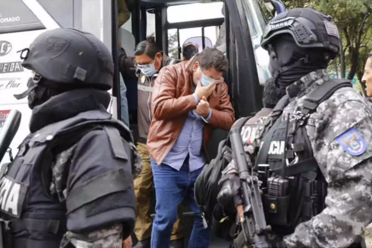 Personas con mascarillas bajando de un autobús escoltadas por policías fuertemente armados.