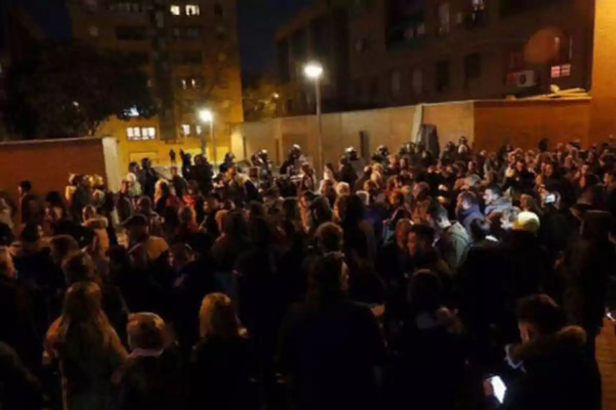 Una multitud de personas se reúne en una calle por la noche, con edificios y luces de fondo.