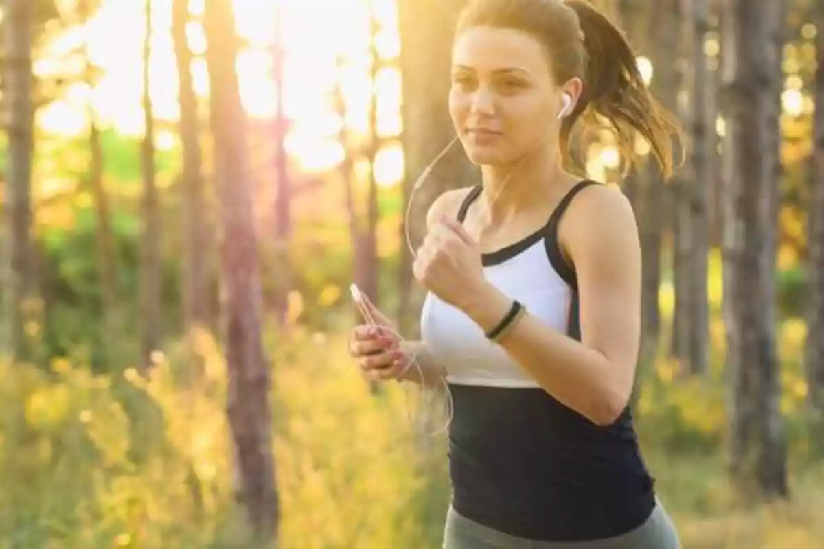 Mujer corriendo en un bosque con auriculares y sosteniendo un teléfono móvil.