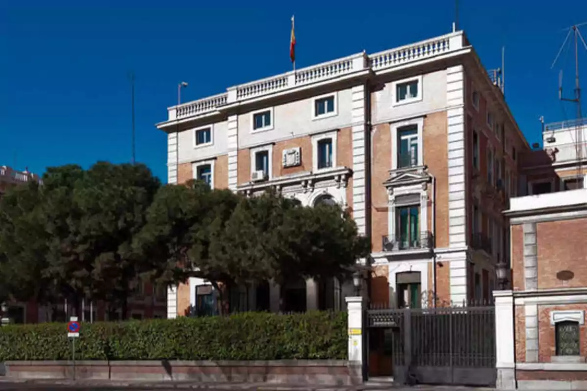 Edificio de ladrillo con detalles blancos y una bandera en la parte superior, rodeado de árboles y una cerca de hierro.