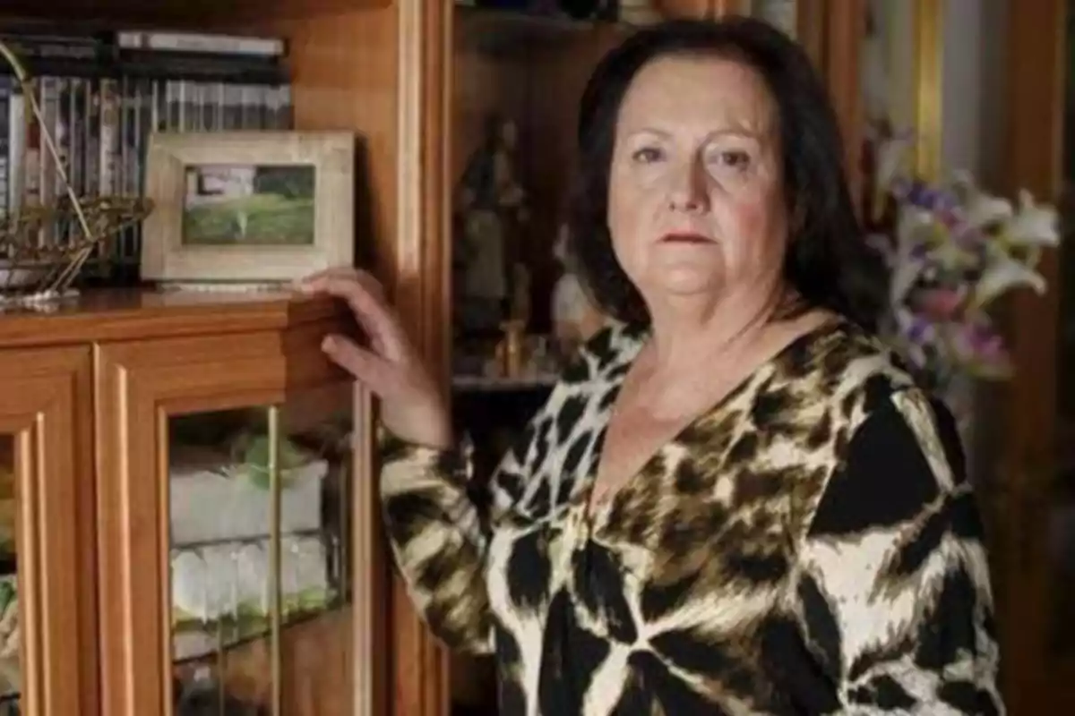 Una mujer de pie junto a un mueble de madera con puertas de vidrio, sosteniendo un marco de fotos.