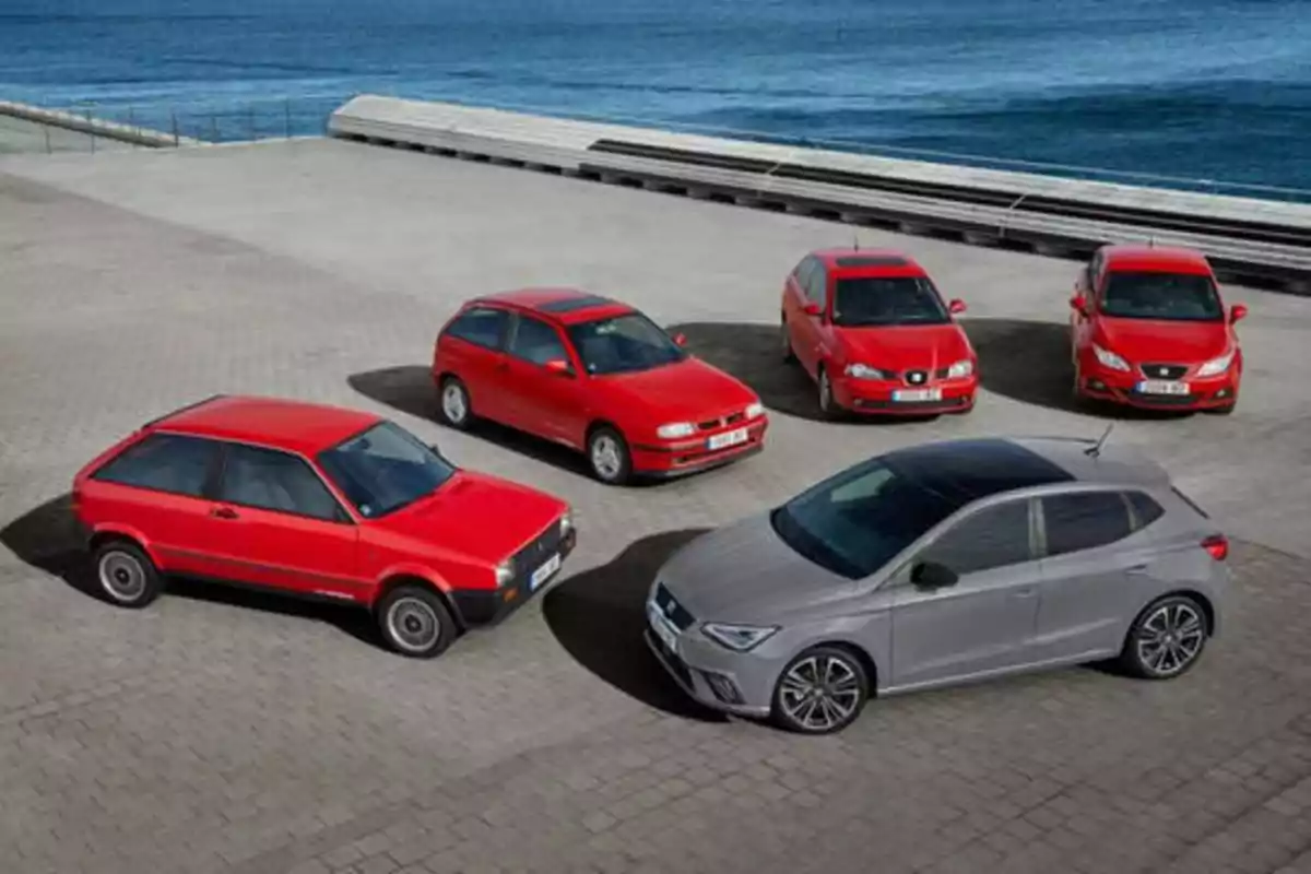 Cinco autos rojos y uno gris estacionados en un área pavimentada cerca del mar.