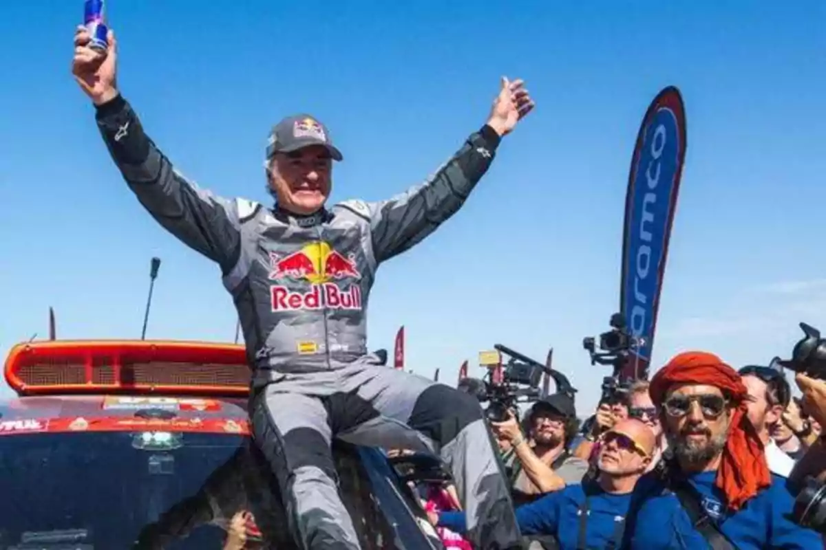Un piloto de carreras celebra su victoria levantando los brazos mientras está sentado en su vehículo rodeado de fotógrafos y aficionados.