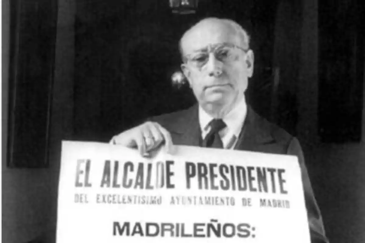 Un hombre mayor con gafas y traje sostiene un cartel que dice "El Alcalde Presidente del Excelentísimo Ayuntamiento de Madrid Madrileños".