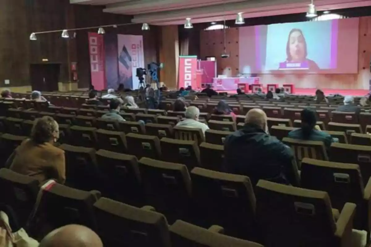 Una conferencia en un auditorio con varias personas sentadas y una pantalla grande al frente mostrando a una persona hablando.