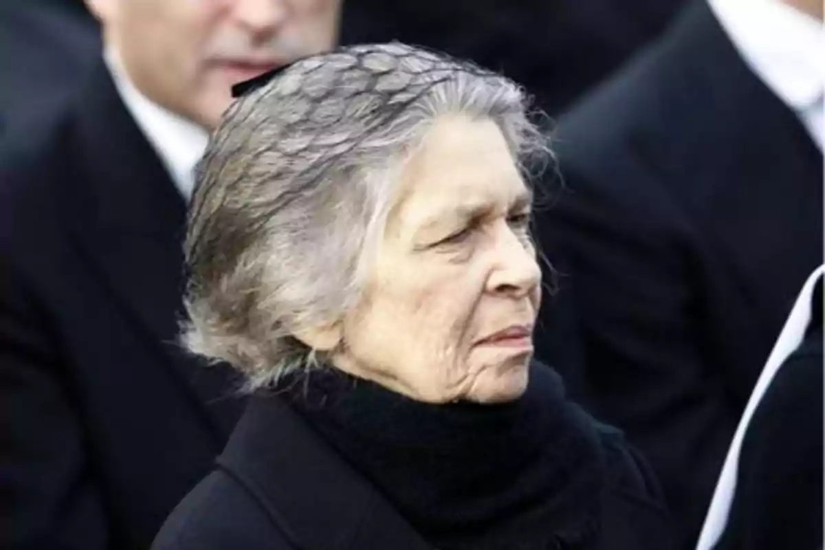 Una mujer mayor con cabello canoso y expresión seria, vestida de negro y con un velo en la cabeza, en un evento formal.