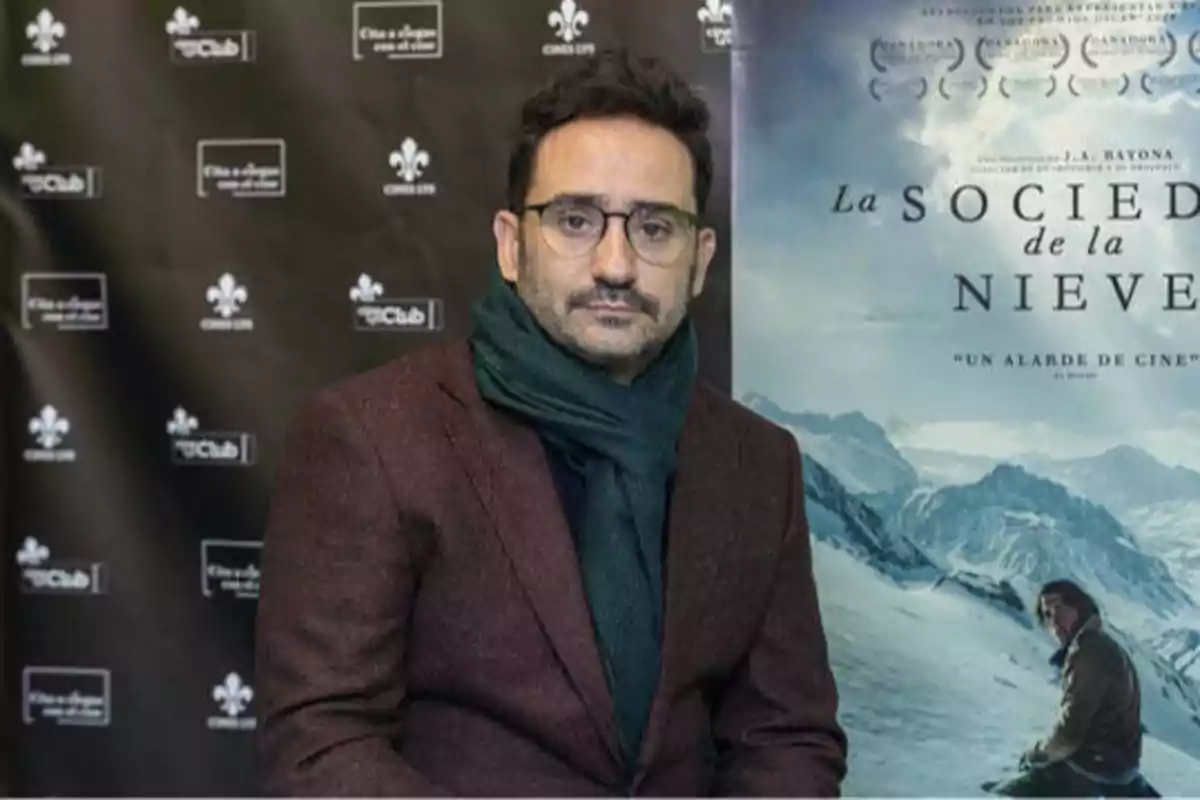 Un hombre con gafas y bufanda posa frente a un cartel de la película "La Sociedad de la Nieve" y un fondo con logotipos de Cine Club.