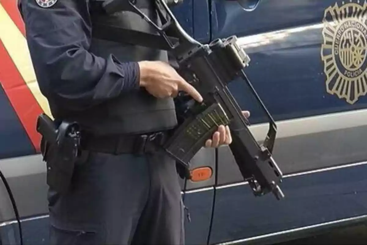 Un policía sosteniendo un arma frente a un vehículo policial.