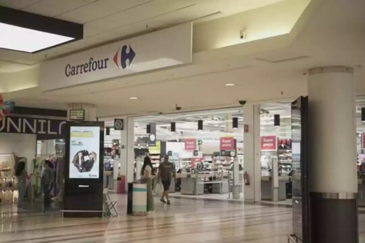 Entrada de un supermercado Carrefour en un centro comercial, con personas caminando y un cartel publicitario en primer plano.