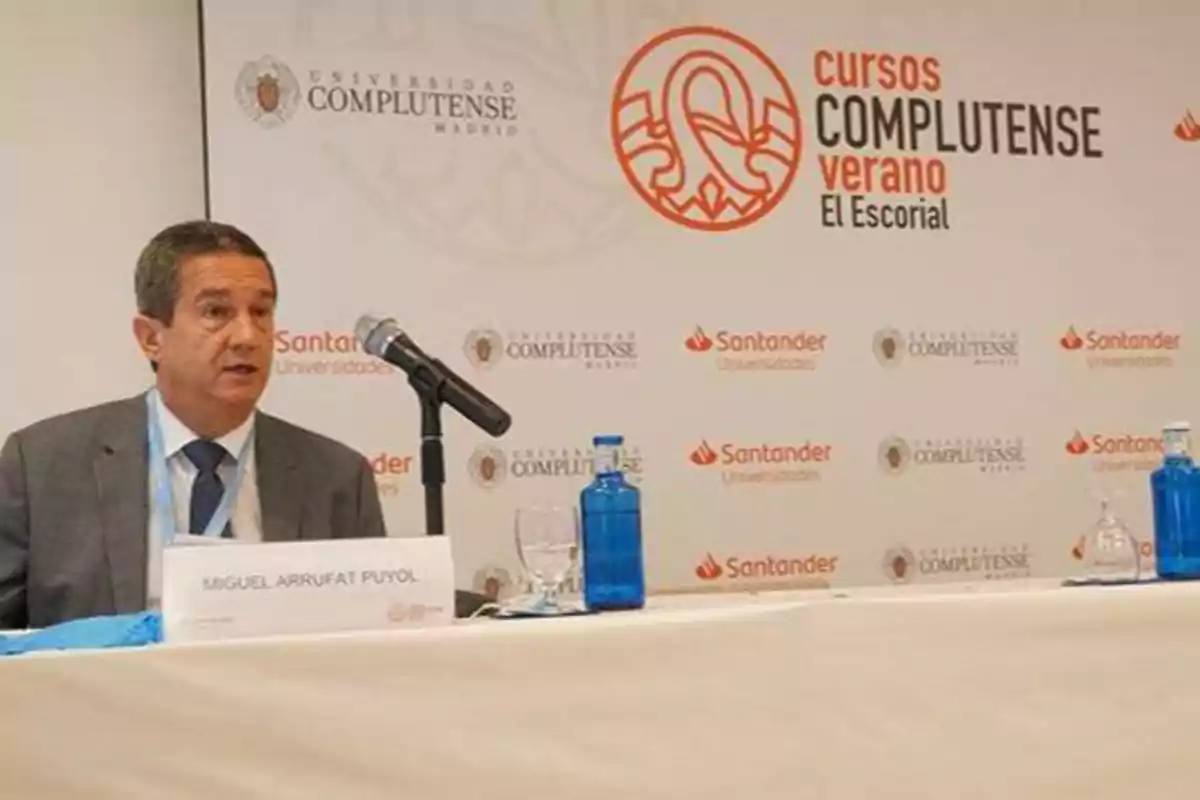 Un hombre está sentado en una mesa con un micrófono frente a él, en un evento de la Universidad Complutense de Madrid, con logotipos de Santander Universidades y Cursos Complutense de Verano El Escorial en el fondo.